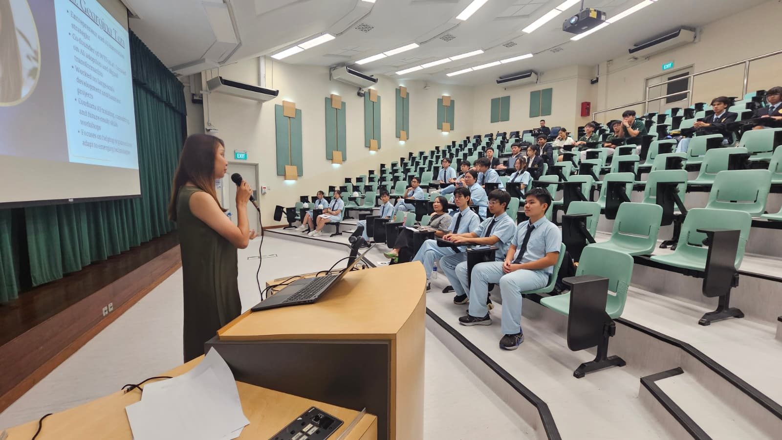 Photograph of Geargina Tan (Coach Gina), Singapore's AI expert and COO of WTFox.ai, on the judging panel in the lecture theatre at Catholic Junior College for Bizcomp 2026, where student teams pitched how AI can help SMEs. Real photo of judging, not a talk.
