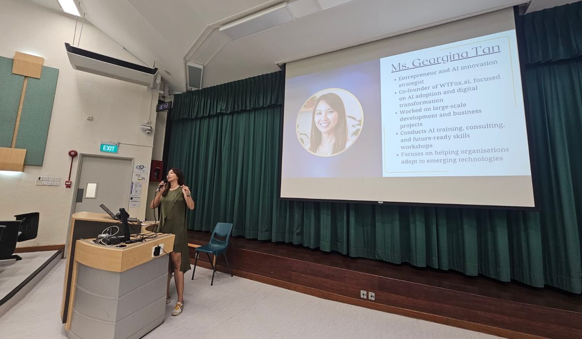 Photograph of Geargina Tan (Coach Gina), Singapore AI speaker and COO of WTFox.ai, presenting at a lectern beside her introduction slide. Real photo. AI speaker Singapore.